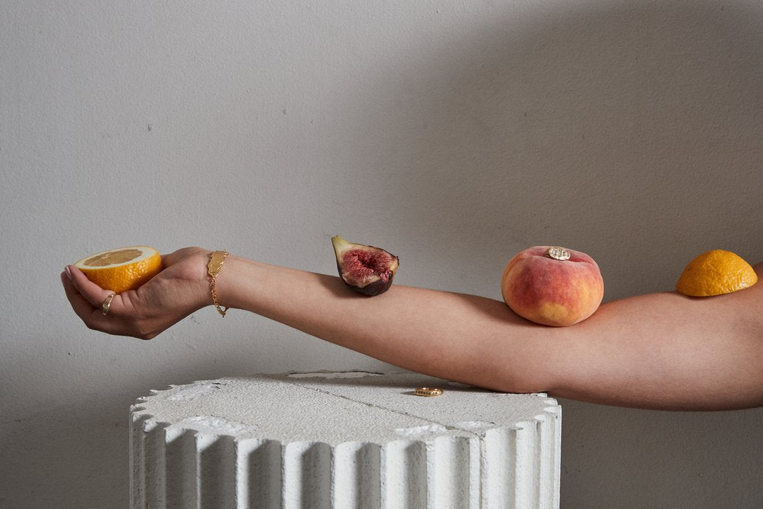 An arm rests on an ancient pillar and holds an orange segment, with a fig, peach and orange balanced on it, wearing decadent jewellery by Gabriella Luchini, including a gold bracelet and gold rings